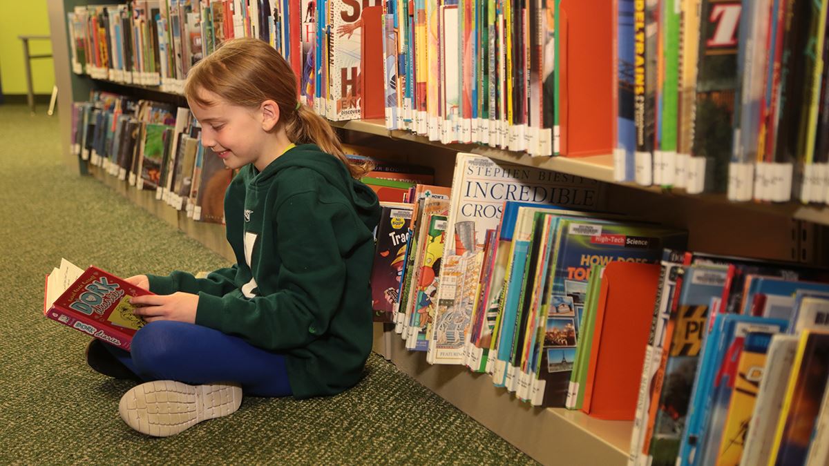 Girl reading a book sitting on the floor in front of a bookshelf